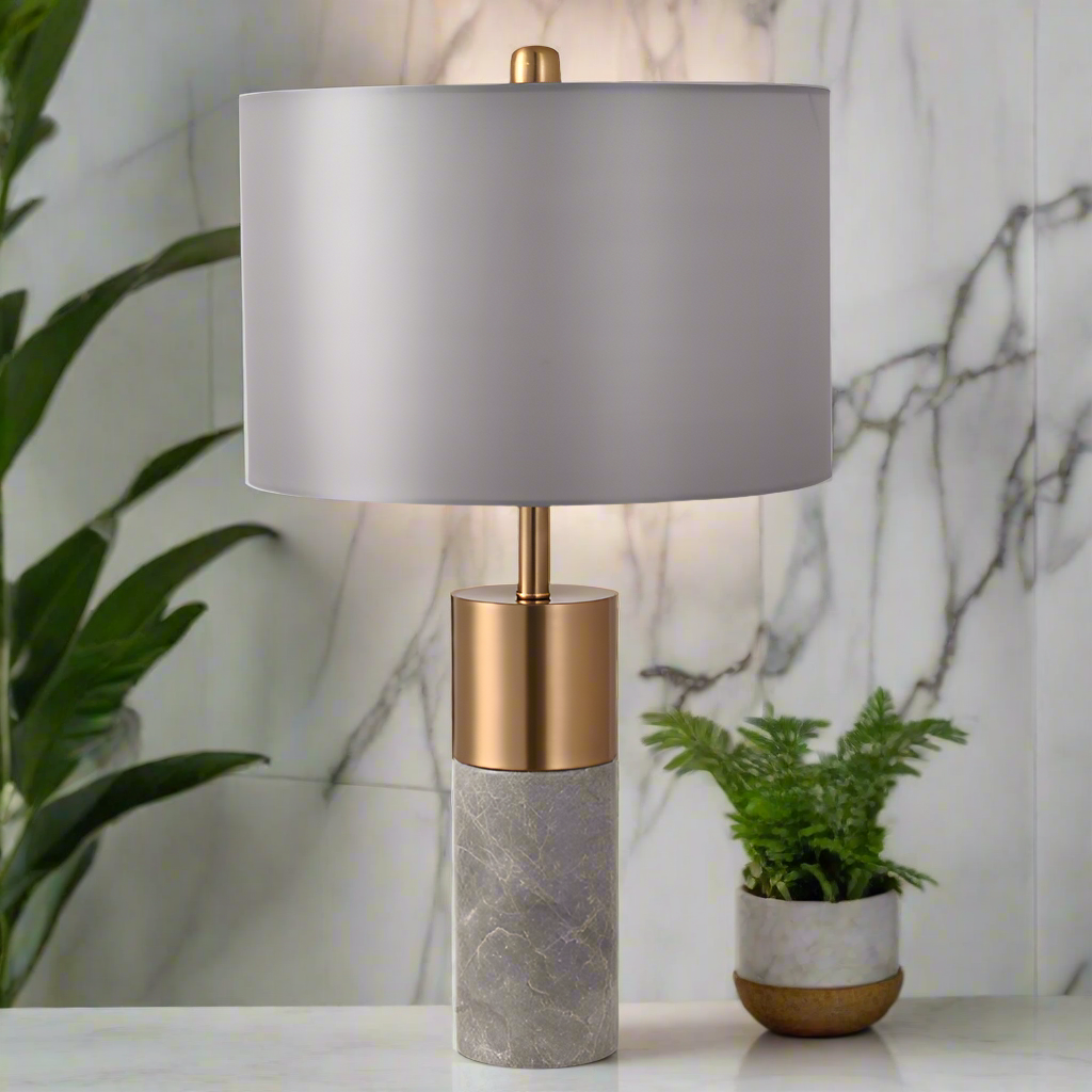 Table lamp with gray shade and gold base on a marble surface with plants in the background